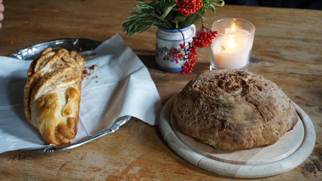 Ein rustikaler Tisch mit zwei Broten, einem geflochtenen Brot auf einem Teller und einem runden Laib auf einem Holzbrett. Neben den Broten steht eine kleine Vase mit frischen Zweigen und einer Kerze. | © Naturpark Sölktäler