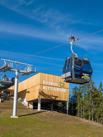 A cable car travels over a green meadow. In the background is the valley station building with trees and blue sky. | © Hans-Peter Steiner