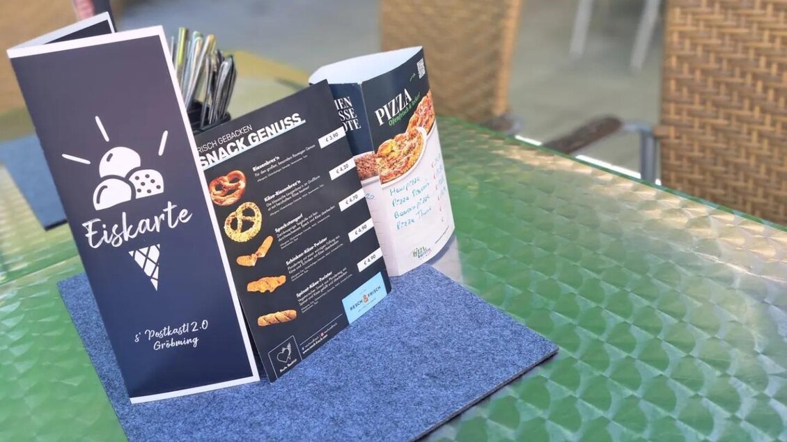 Eine menükarte mit verschiedenen Snacks und einer Eiskarte liegt auf einem Tisch mit einer grünen Glasoberfläche. Im Hintergrund sind Stühle und eine entspannte Außenatmosphäre zu sehen. | © Thomas Bartl