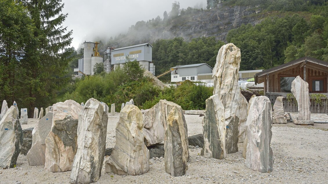 Eine Gruppe von großen, unregelmäßig geformten Steinen steht in einer grauen Umgebung. Im Hintergrund sind Gebäude und bewaldete Hügel zu sehen, während Nebel über der Landschaft hängt. | © Herfried Marek