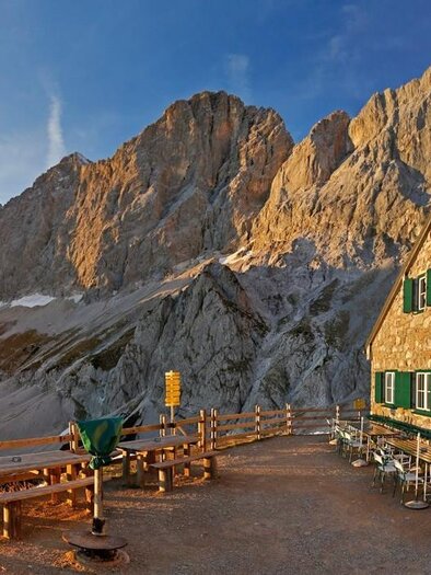 Eine Berglandschaft mit beeindruckenden Felsen und einer Berghütte. Die Hütte hat grüne Fenster und eine Terrasse mit Tischen und Stühlen. | © Herbert Raffalt