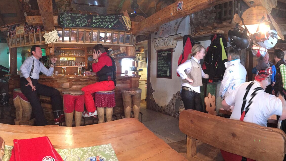 Eine gemütliche Hütte mit Holzdekor, in der Menschen am Tisch sitzen und an der Bar stehen. Die Gäste tragen winterliche Kleidung und scheinen sich zu unterhalten. | © Sautrog-Alm