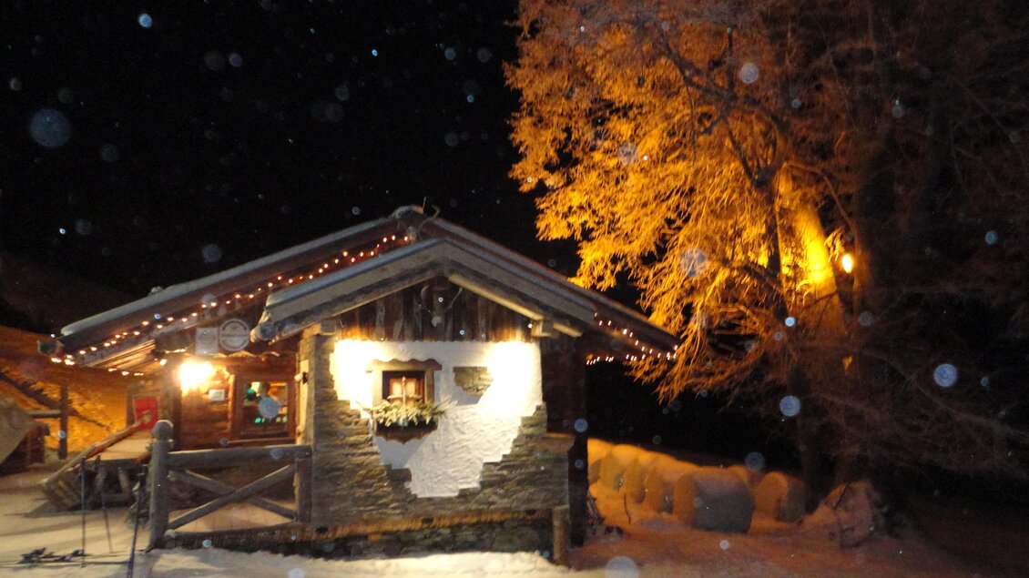Ein gemütliches Holzhaus im Schnee, beleuchtet von winterlichen Lichtern. Ein großer Baum mit goldenem Licht steht daneben. | © Sautrog-Alm