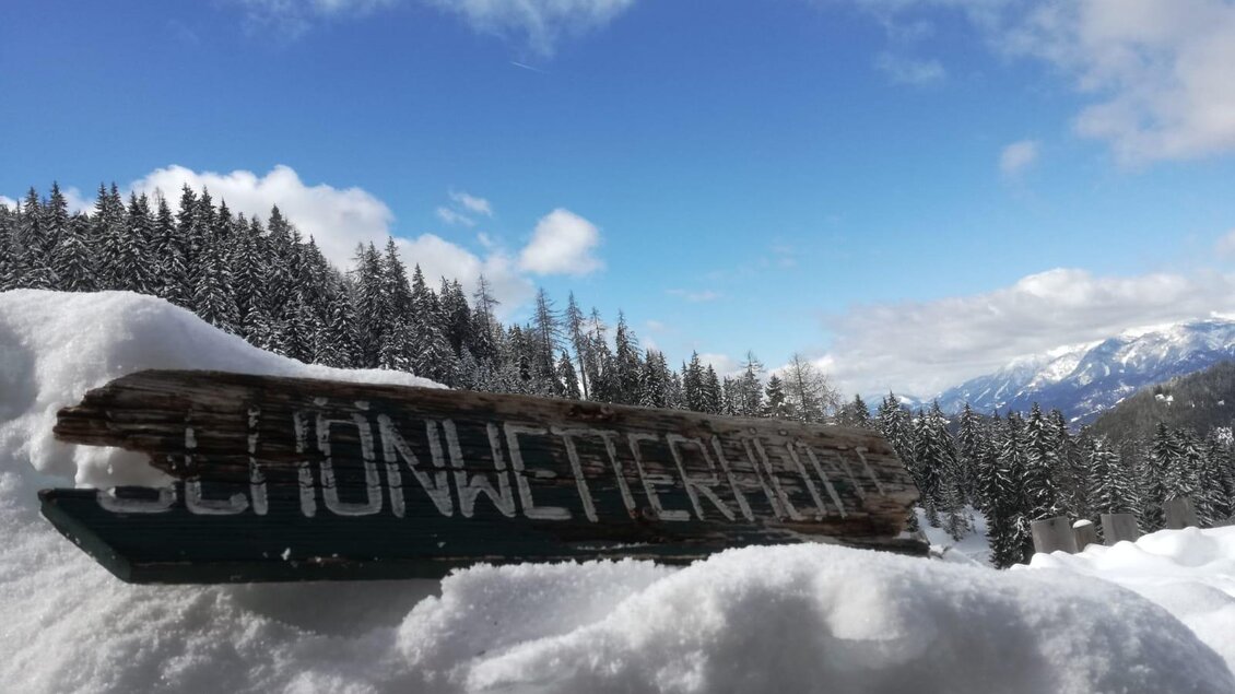 Ein Holzschild mit der Aufschrift „Schönwetterhöhe“ steht im Schnee. Im Hintergrund sind schneebedeckte Berge und ein blauer Himmel zu sehen. | © Familie Brugger
