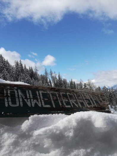 A wooden sign with the inscription "Schönwetterhöhe" stands in the snow. In the background, snow-covered mountains and a blue sky can be seen. | © Familie Brugger