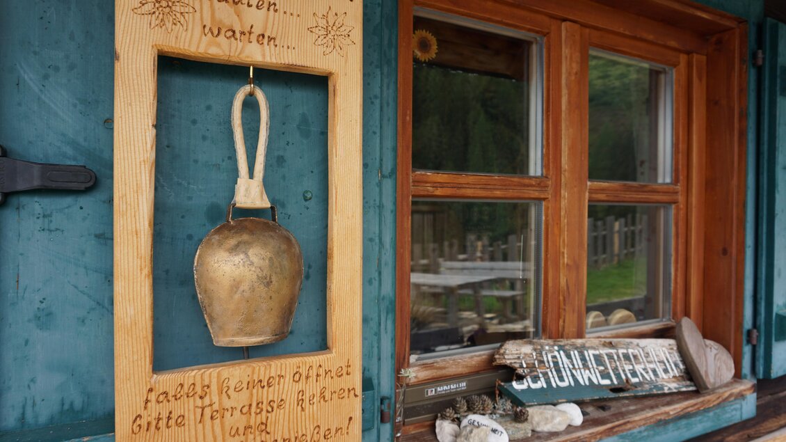 Ein rustikaler Holzrahmen mit einer Glocke und einem einladenden Schild hängt an einem Fenster. Das Schild fordert dazu auf, die Terrasse zu betreten und zu genießen. | © Naturpark Sölktäler