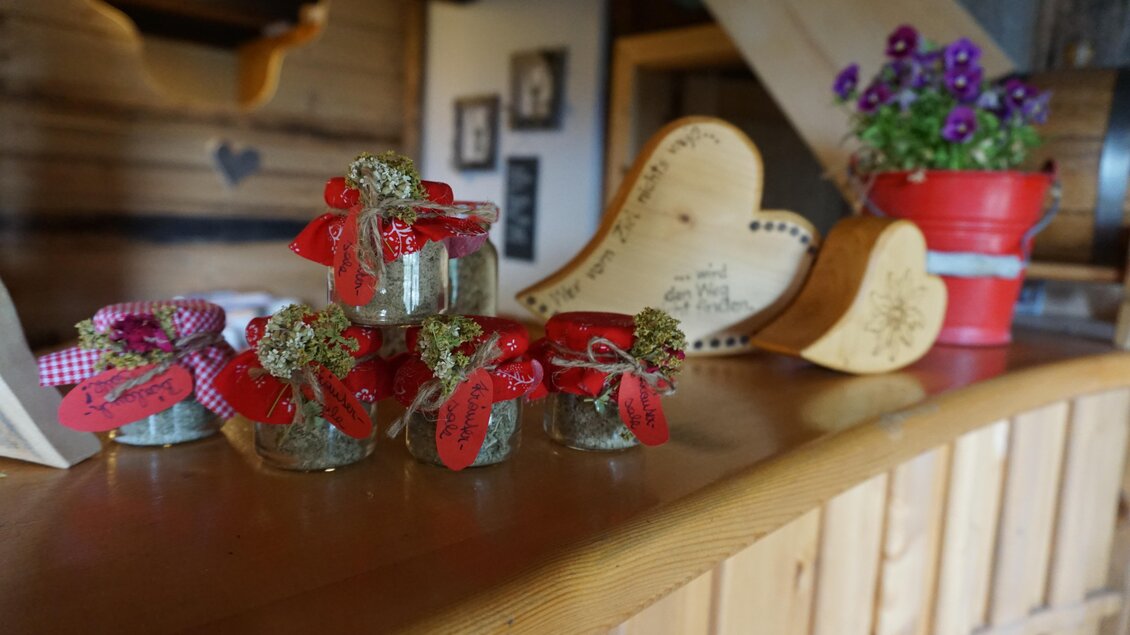 Ein rustikaler Tisch mit kleinen Gläschen, die mit Kräutern gefüllt sind und rot-weiß karierten Bändern dekoriert sind. Daneben steht eine Blume in einem Topf und ein herzförmiges Holzschild. | © Naturpark Sölktäler