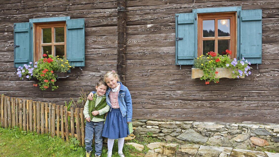 Zwei Kinder posieren fröhlich vor einer Holzfassade mit blauen Fensterläden. Vor den Fenstern blühen bunte Blumen in Kästen. | © Herfried Marek