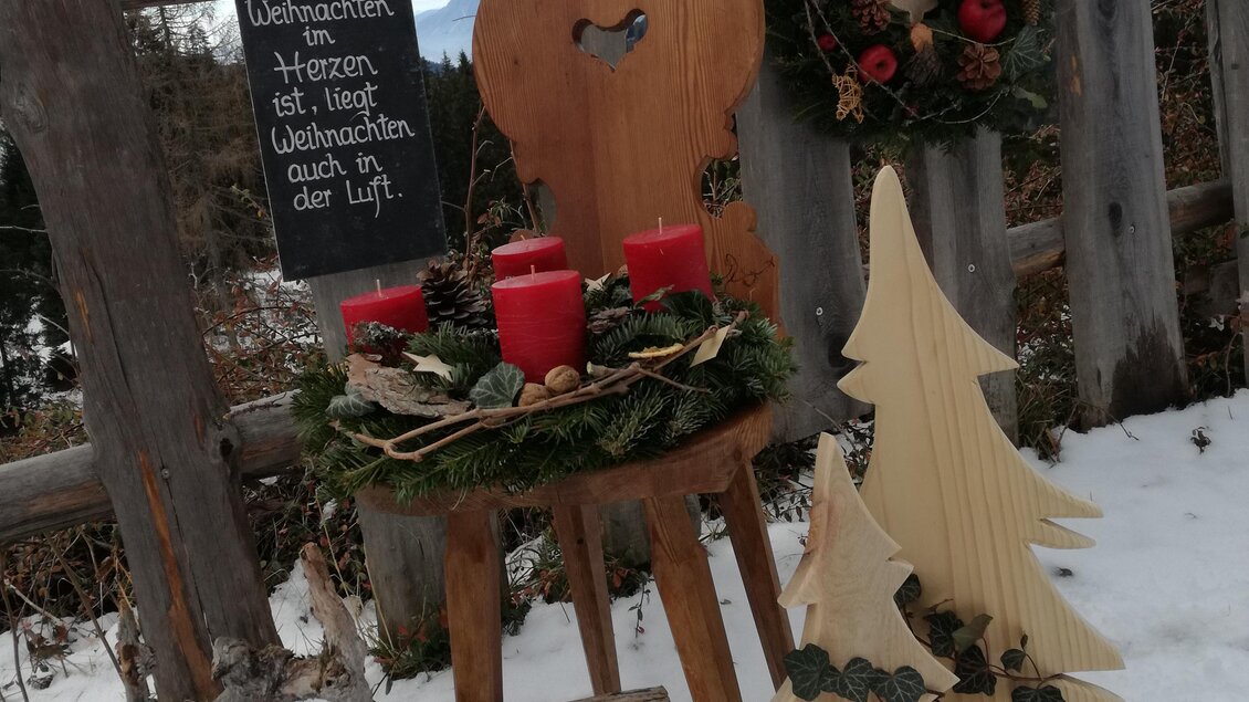 Eine rustikale Weihnachtsdekoration im Schnee mit einem Holzstuhl, roten Kerzen und einem Kranz. Neben dem Stuhl steht ein dekorativer Holzbaum und ein Schild mit der Aufschrift „Schönwetterheit“. | © Familie Brugger