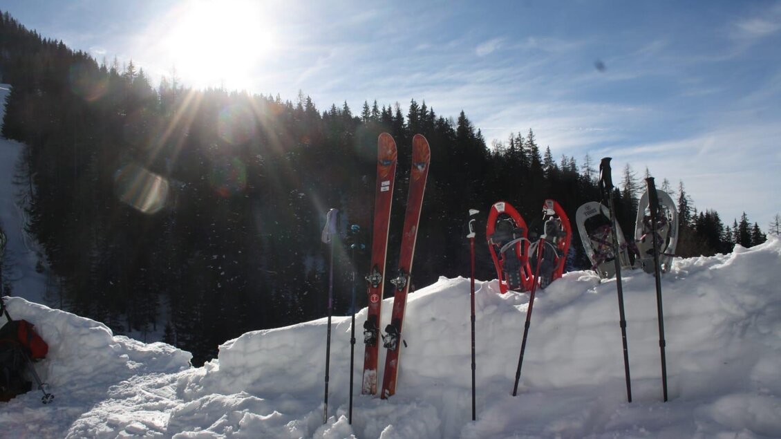 Eine verschneite Berglandschaft mit Skiern und Schneeschuhen steht in der Sonne. Im Hintergrund sind Bäume und ein klarer Himmel zu sehen. | © Familie Brugger