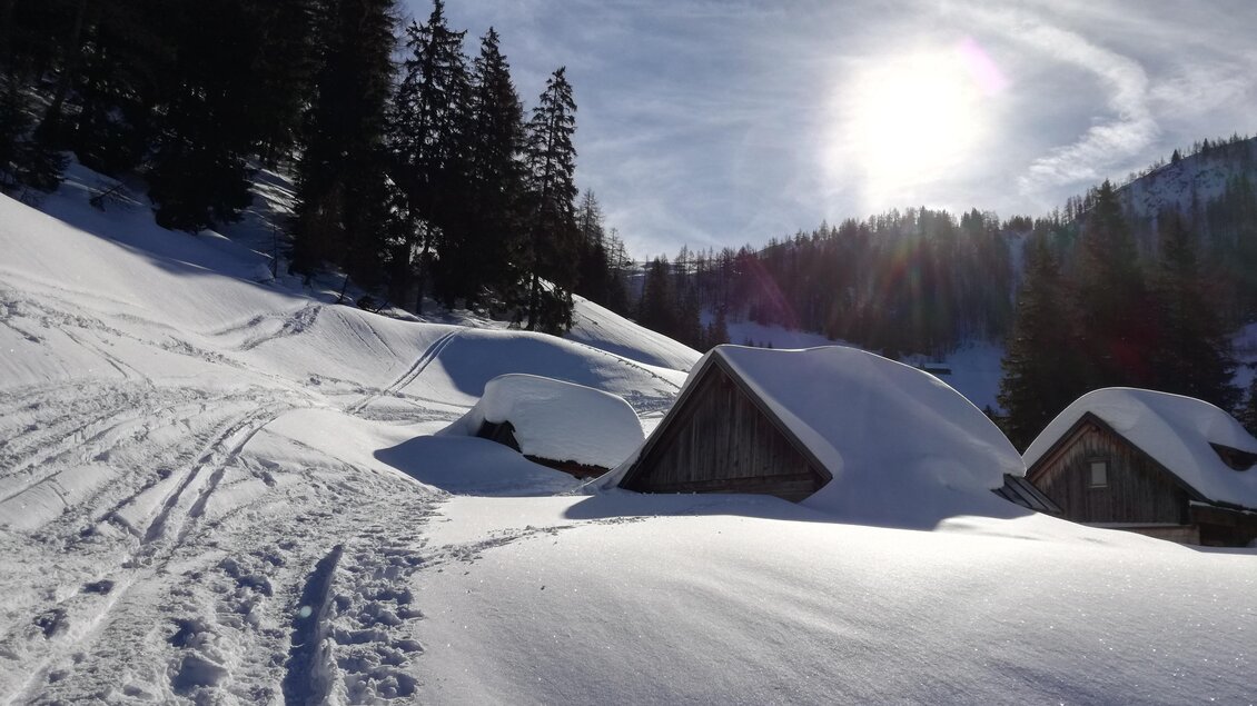 Eine winterliche Landschaft mit schneebedeckten Hütten und hohem Schnee. Die Sonne scheint durch den klaren Himmel und beleuchtet die Bäume im Hintergrund. | © Familie Brugger