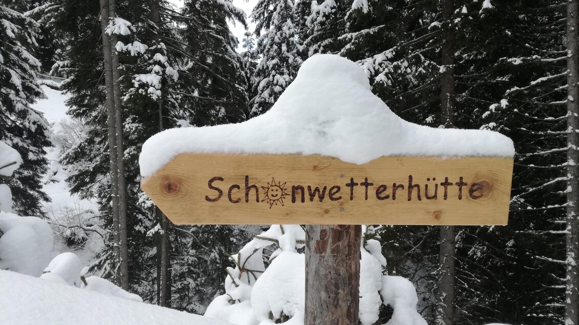 Ein Holzschild mit der Aufschrift „Schönwetterhütte“ steht im Schnee. Umgeben von verschneiten Bäumen vermittelt die Szene eine ruhige Winteratmosphäre. | © Familie Brugger