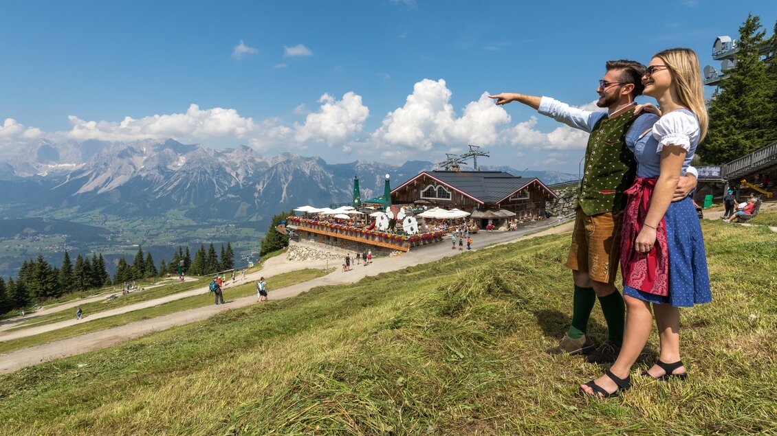 Ein Paar in traditioneller Kleidung steht auf einer Wiese und zeigt auf die Berge im Hintergrund. Im Vordergrund sind blühende Landschaften und ein Bergrestaurant zu sehen. | © Harald Steiner