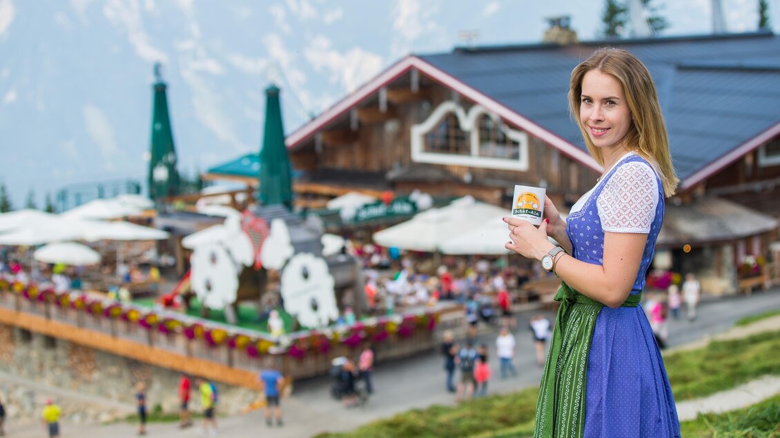Eine Frau in traditioneller Tracht steht vor einem bayerischen Gasthaus in den Bergen. Im Hintergrund sind viele Gäste und die schöne Landschaft zu sehen. | © Harald Steiner