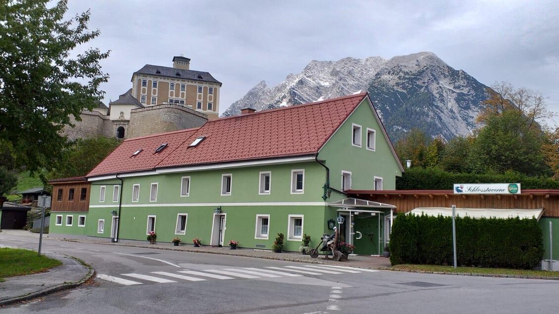 Ein grünes Gebäude mit rotem Dach befindet sich an einer Straßenkreuzung. Im Hintergrund sind die Berge und ein bewölkter Himmel zu sehen. | © Schlosstaverne