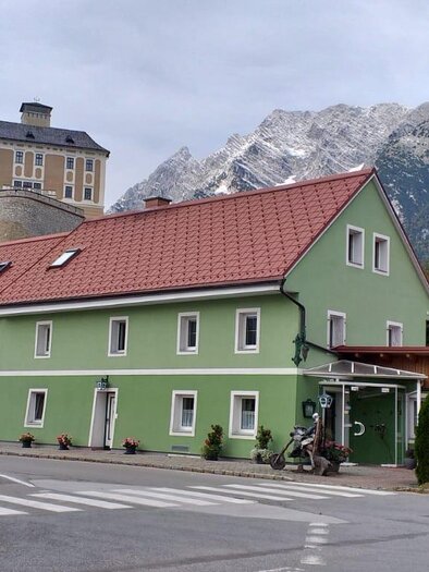 Ein grünes Gebäude mit rotem Dach befindet sich an einer Straßenkreuzung. Im Hintergrund sind die Berge und ein bewölkter Himmel zu sehen. | © Schlosstaverne