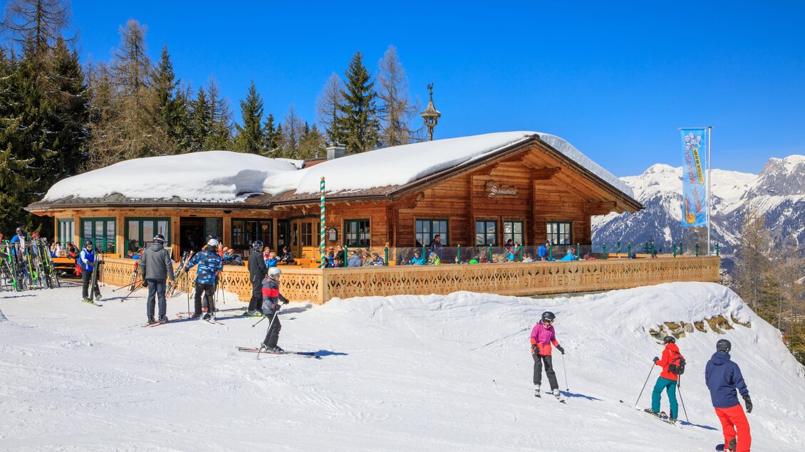 Eine gemütliche Skihütte umgeben von schneebedeckten Bergen. Skifahrer genießen den sonnigen Tag auf der Piste. | © Schmiedhütte