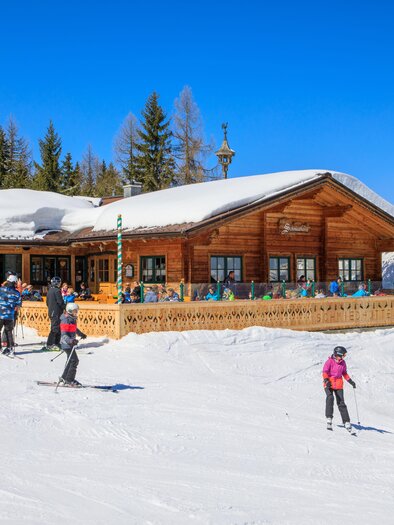 A cozy ski lodge surrounded by snow-covered mountains. Skiers are enjoying the sunny day on the slopes. | © Schmiedhütte
