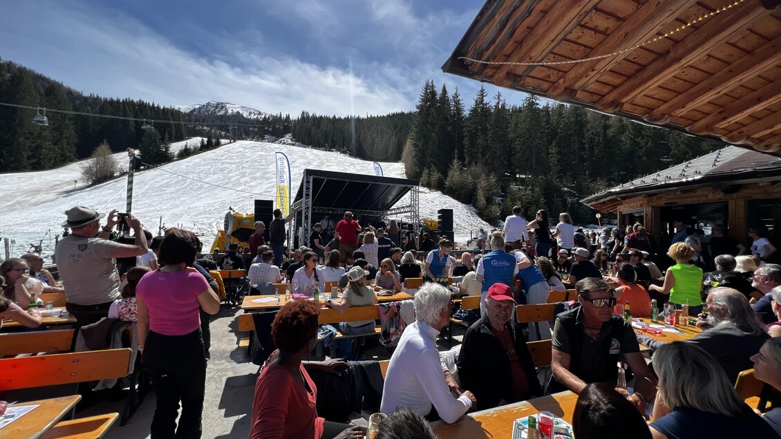 Ein lebhaftes Fest im Freien mit vielen Menschen, die an Tischen sitzen. Im Hintergrund sind schneebedeckte Berge und eine Bühne für Live-Musik zu sehen. | © Schmiedhütte