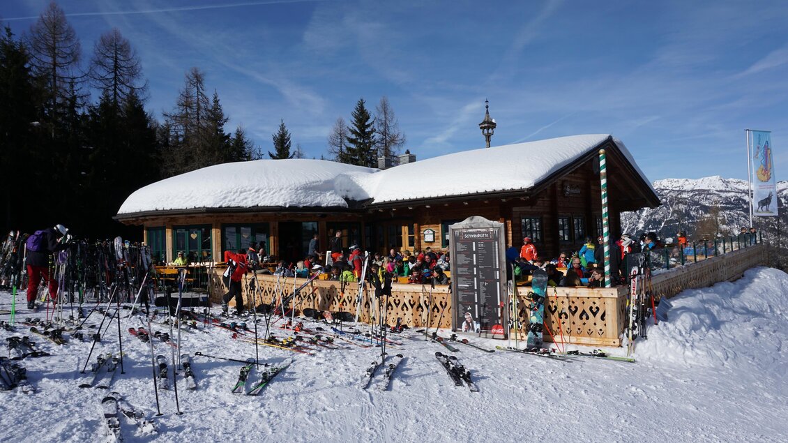 Eine gemütliche Berghütte im Schnee mit vielen Skifahrern und Gästen. Die Umgebung ist von verschneiten Bäumen und klarem Himmel geprägt. | © Schmiedhütte