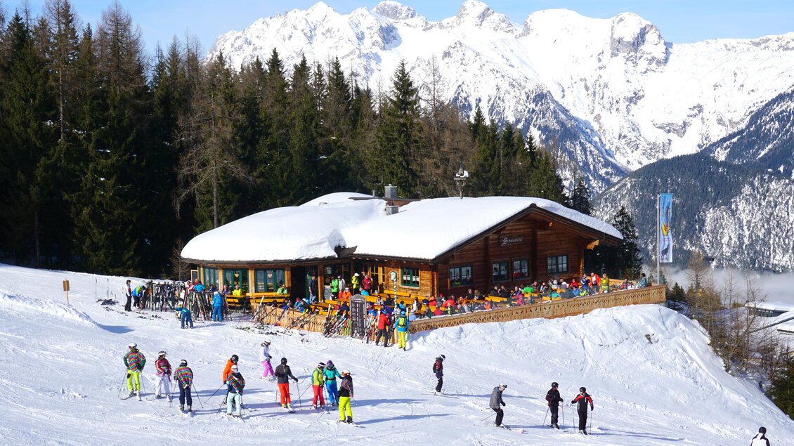 Eine gemütliche Skihütte in den Bergen, umgeben von Schnee und Bäumen. Viele Skifahrer genießen die Aussicht und die Sonne. | © Schmiedhütte