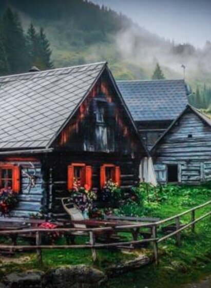 A charming mountain cabin with colorful flowers and an inviting terrace. Surrounded by green forests and light mist. | © Schrabachalm
