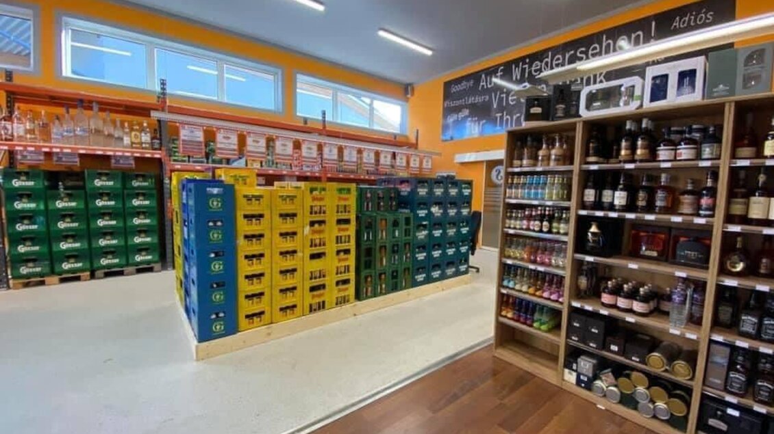 Ein gut sortierter Verkaufsraum mit Regalen voller Getränke und Lebensmitteln. Die Wände sind orange gestrichen und große Fenster sorgen für Tageslicht. | © Schrotti's Getränkeshop