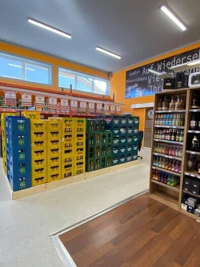 A well-organized sales area with shelves full of drinks and groceries. The walls are painted orange, and large windows provide natural light. | © Schrotti's Getränkeshop