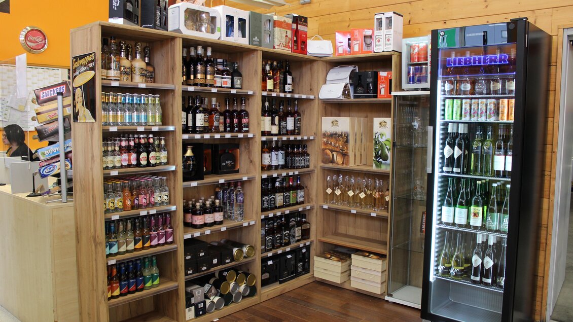 Ein Verkaufsregal mit einer Vielzahl von Getränken, darunter alkoholische und nicht-alkoholische Optionen. Neben dem Regal steht ein Kühlschrank mit Flaschen und Gläsern. | © Schrotti's Getränkeshop