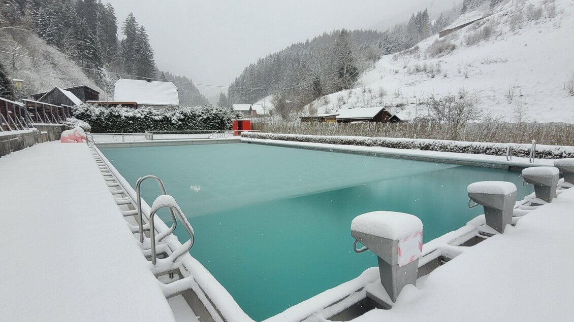 Ein leeres Schwimmbad im Schnee umgeben von Bergen und Hütten. Die Wasseroberfläche ist grün und liegt unter einer Schicht aus frischem Schnee. | © Tim Fuchsberger