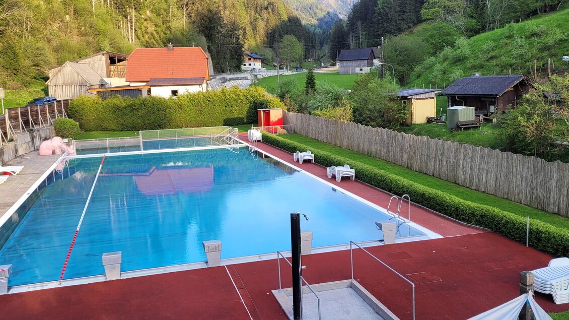 Ein hübscher Swimmingpool umgeben von grünen Wäldern und gemütlichen Holzhäusern. Entspannte Liegen laden zum Verweilen ein. | © Tim Fuchsberger