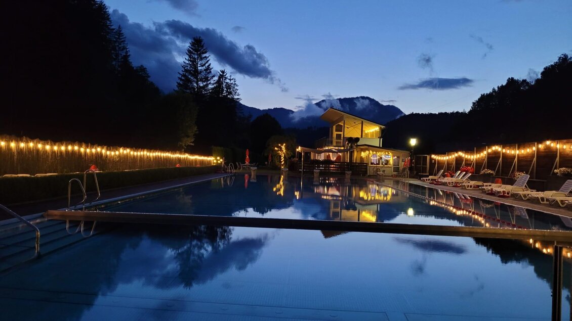 Ein schöner Pool bei Nacht, umgeben von Lichtern und Bergen. Das Wasser spiegelt die Umgebung und schafft eine entspannte Atmosphäre. | © Tim Fuchsberger