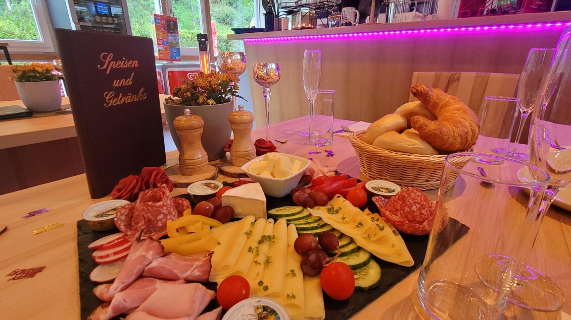 Ein schön angerichteter Teller mit verschiedenen Wurst- und Käsesorten, frischem Gemüse und Trauben. Im Hintergrund sind Gläser und ein Korb mit Brötchen zu sehen. | © Tim Fuchsberger