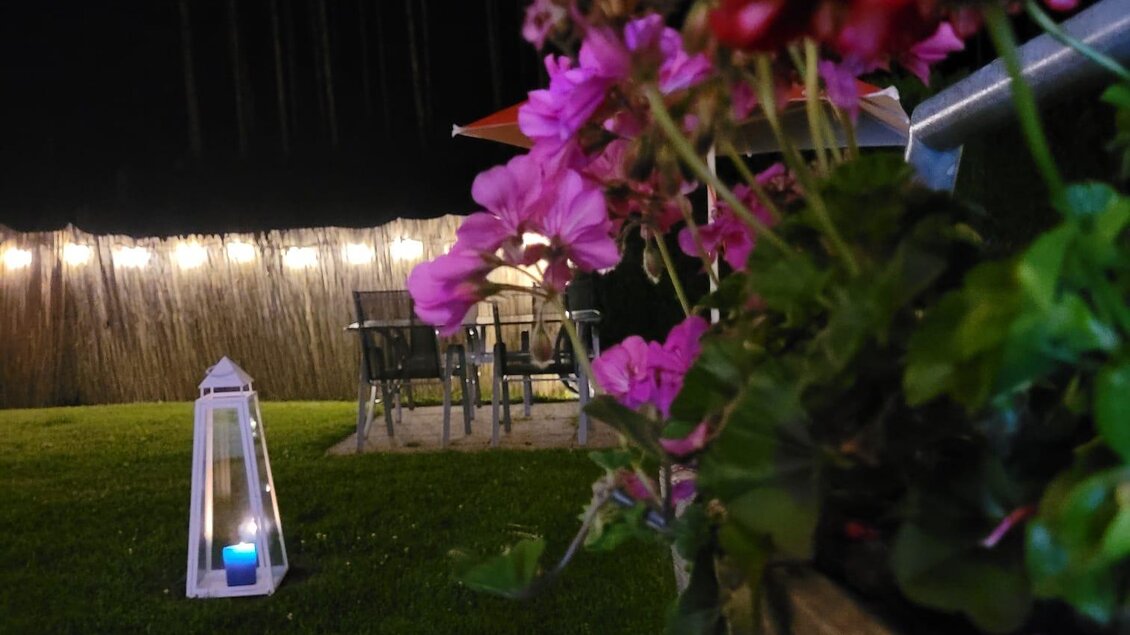 Eine gemütliche Gartenszene bei Nacht mit bunten Blumen im Vordergrund. Im Hintergrund sind ein paar Gartenstühle und eine sanfte Beleuchtung zu sehen. | © Tim Fuchsberger