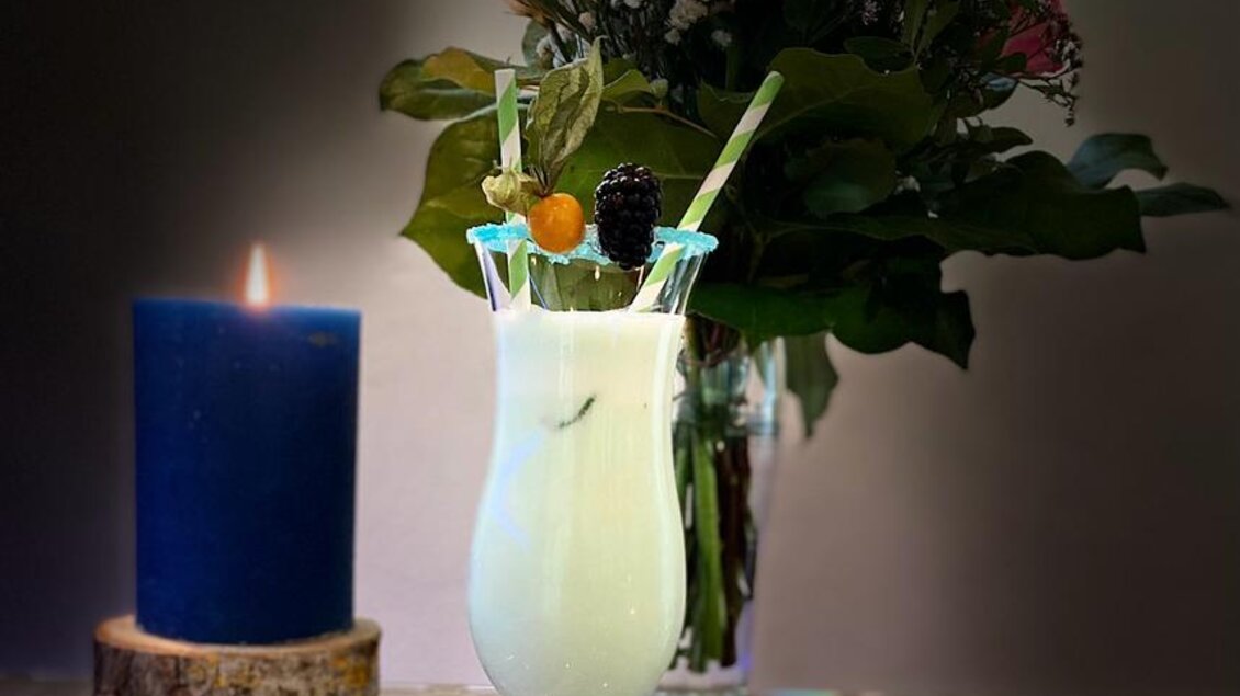 Ein erfrischender Cocktail in einem eleganten Glas mit einer bunten Dekoration. Daneben steht eine blühende Vase und eine brennende Kerze auf einem rustikalen Holzuntersetzer. | © Tim Fuchsberger