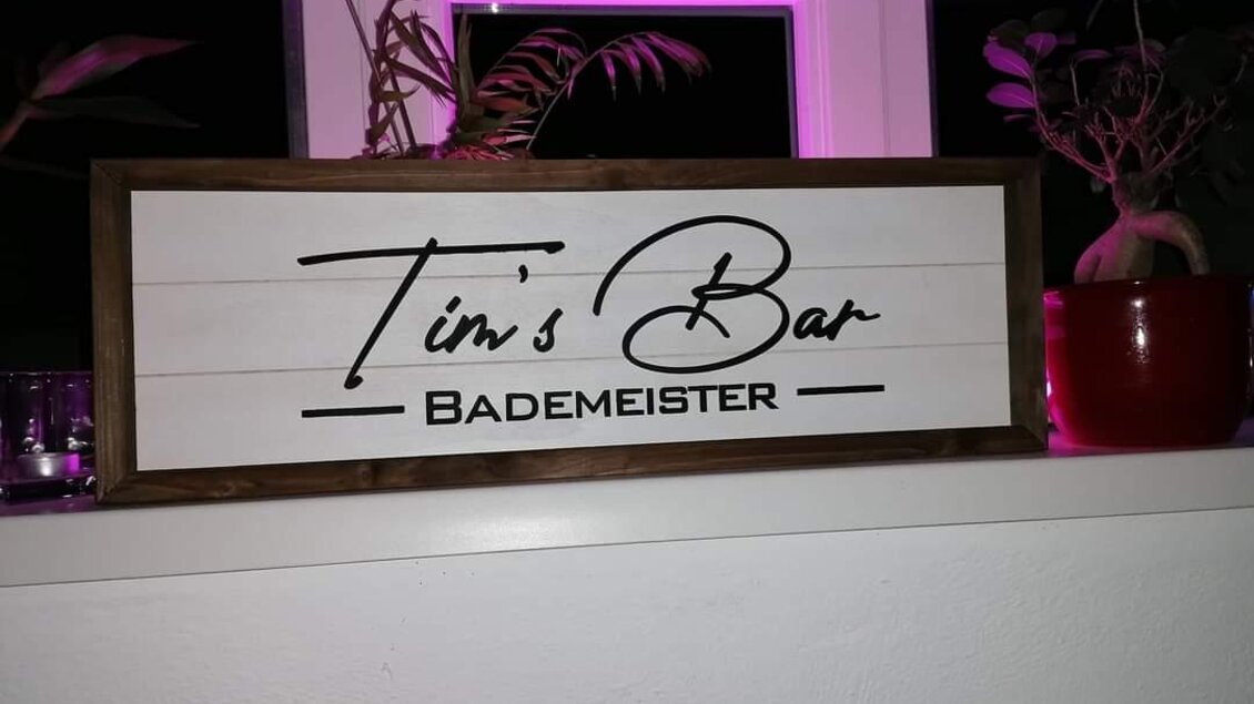 Ein dekoratives Schild mit der Aufschrift "Tim's Bar" und dem Wort "Bademeister". Das Schild ist auf einem Fensterbrett platziert, umgeben von Pflanzen. | © Tim Fuchsberger