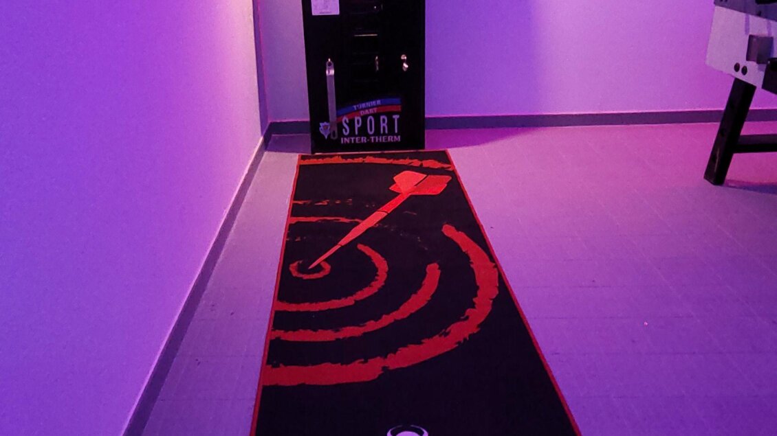 Eine Dartscheibe mit einem dazugehörigen Teppich in einem beleuchteten Raum. Die Wände sind in sanften Lila- und Blautönen gehalten. | © Tim Fuchsberger