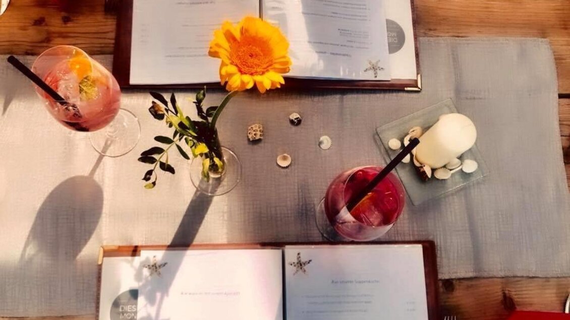Ein gedeckter Tisch mit Speisekarten, zwei Cocktails und einer gelben Blume. Die Atmosphäre ist einladend und entspannt. | © Seestüberl Aich