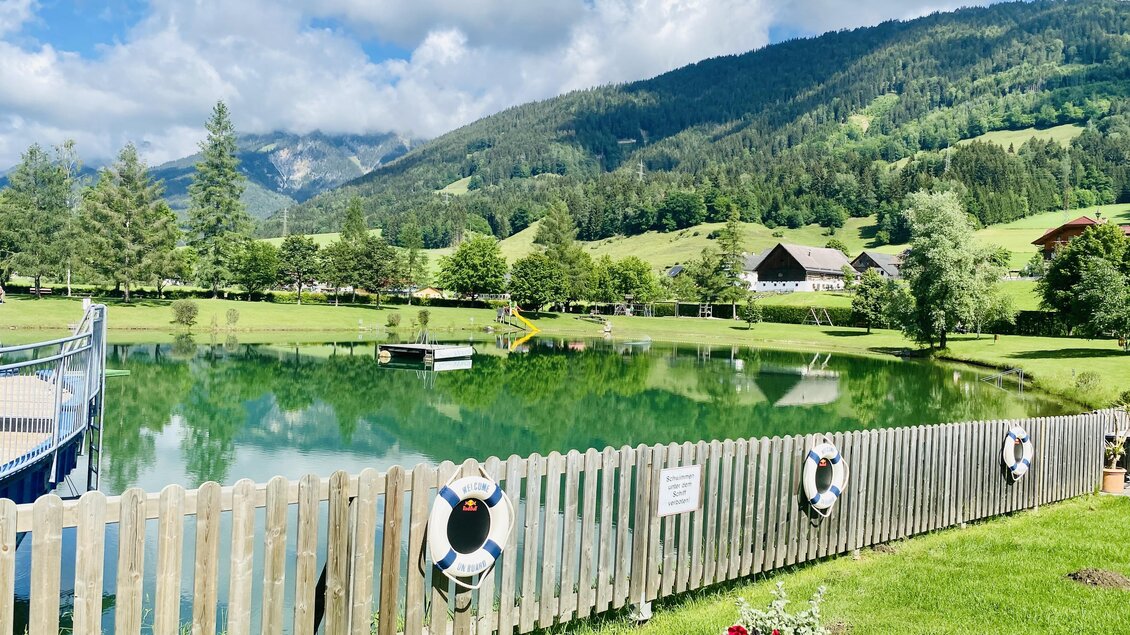 Ein malerischer See, umgeben von grünen Wiesen und Bergen. Im Vordergrund sind Schwimmringe an einem Holzzaun sichtbar. | © Seestüberl Aich