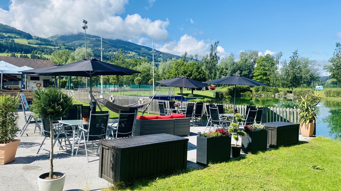 Ein gemütlicher Außenbereich mit Tischen und Stühlen, umgeben von grüner Natur. Im Hintergrund sind Hügel und ein ruhiger See sichtbar. | © Seestüberl Aich