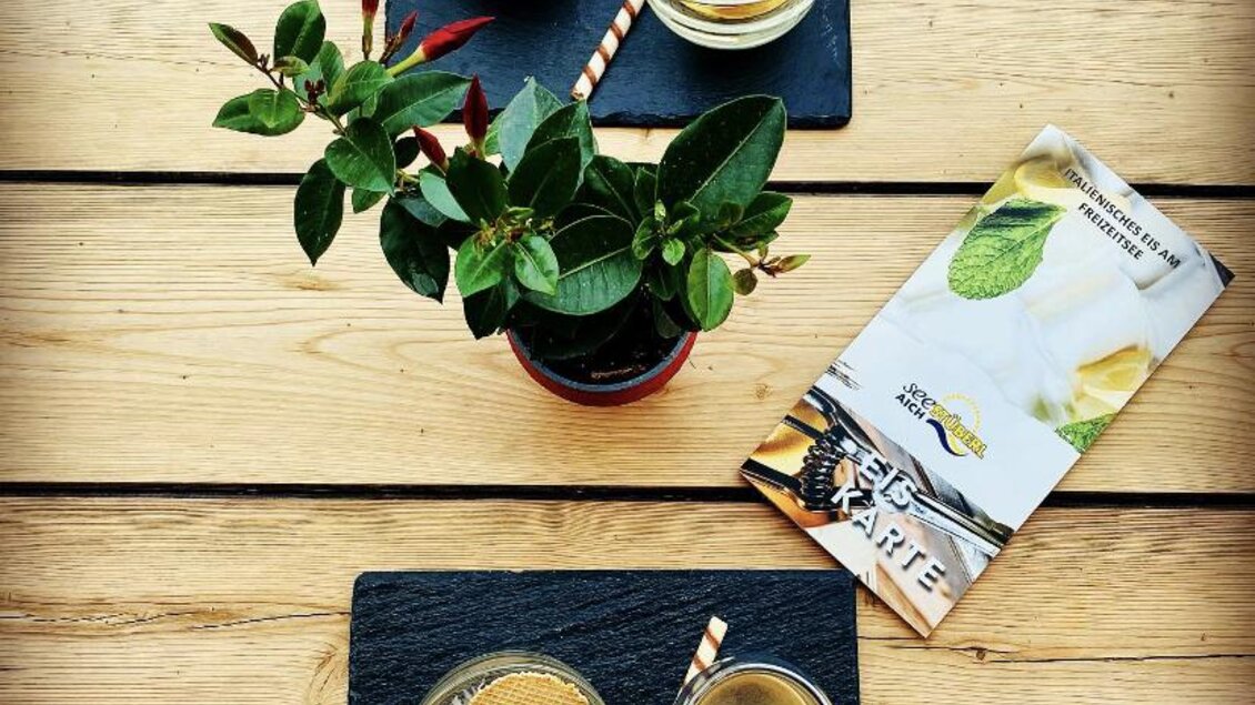 Zwei Gläser mit Getränken und einer Dessertschale auf dunklen Platten. Eine kleine Pflanze steht in der Mitte, dazu ein Faltblatt. | © Seestüberl Aich