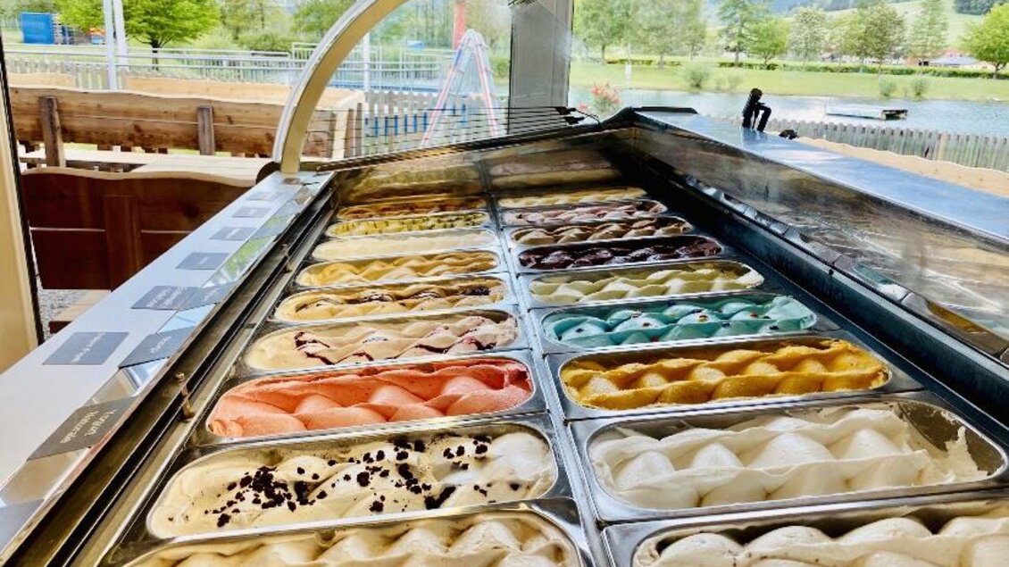 Eine Eisdiele mit vielen bunten Eisvariationen in der Vitrine. Im Hintergrund sind grüne Bäume und eine Spielplatzstruktur zu sehen. | © Seestüberl Aich