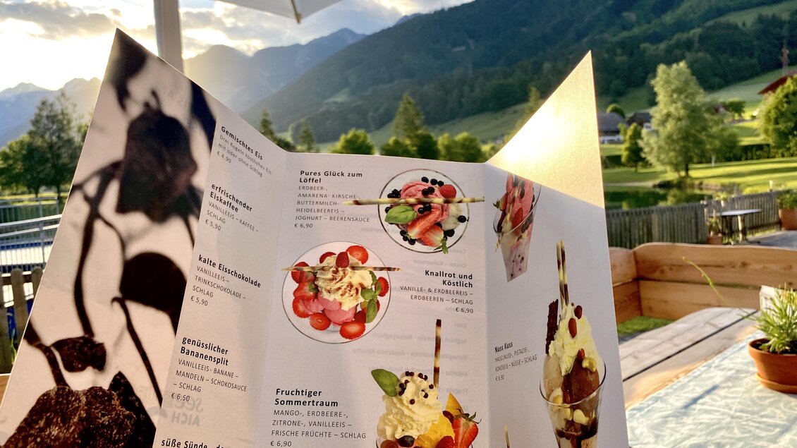 Eine Speisekarte mit verschiedenen Eissorten und frischen Zutaten. Im Hintergrund sind grüne Hügel und ein klarer Himmel zu sehen. | © Seestüberl Aich