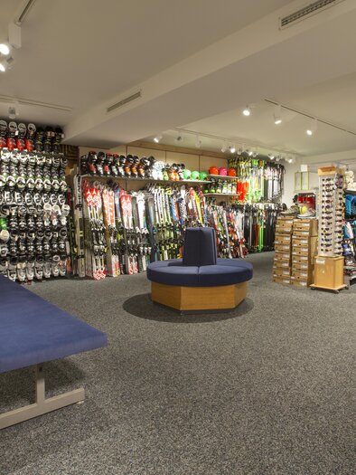 A ski shop with numerous skis, snowboards, and accessories. In the foreground, there is a blue bench in the middle of the room. | © Ski Lenz