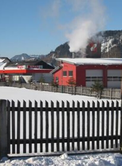 A winter landscape with snow-covered fields and houses in the background. Steam rises from a building and the landscape is framed by mountains.
