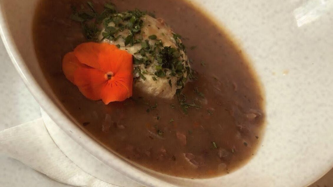 Eine Schüssel mit einer herzhaften Suppe und einer grünen Knödel in der Mitte. Eine Blüte in leuchtendem Orange rundet das Gericht ab. | © Spechtenseehütte