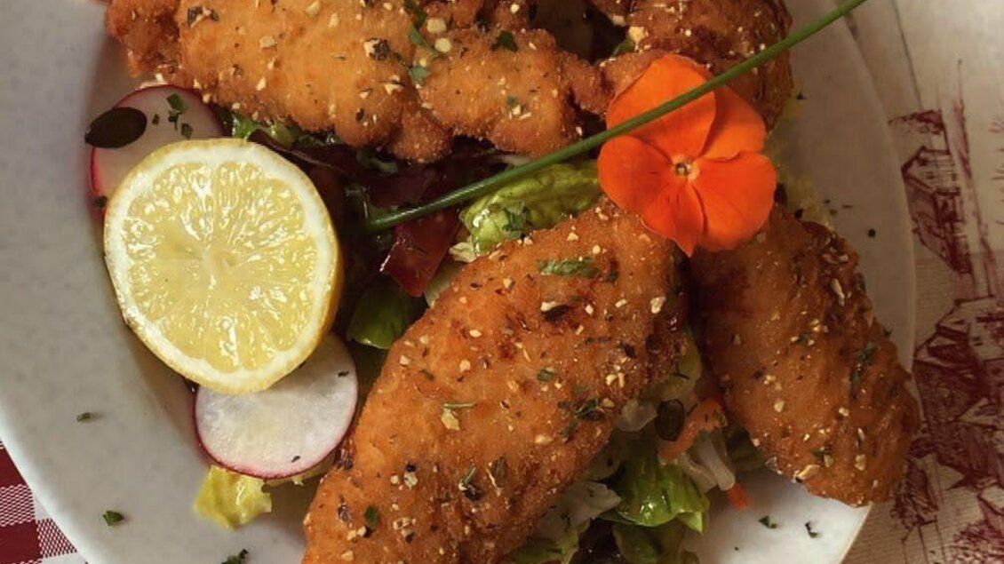 Ein Teller mit frittiertem Fisch und einer Beilage aus frischem Salat. Dazu sind eine Limettenscheibe und eine kleine Garnierung aus Blumen auf dem Gericht. | © Spechtenseehütte