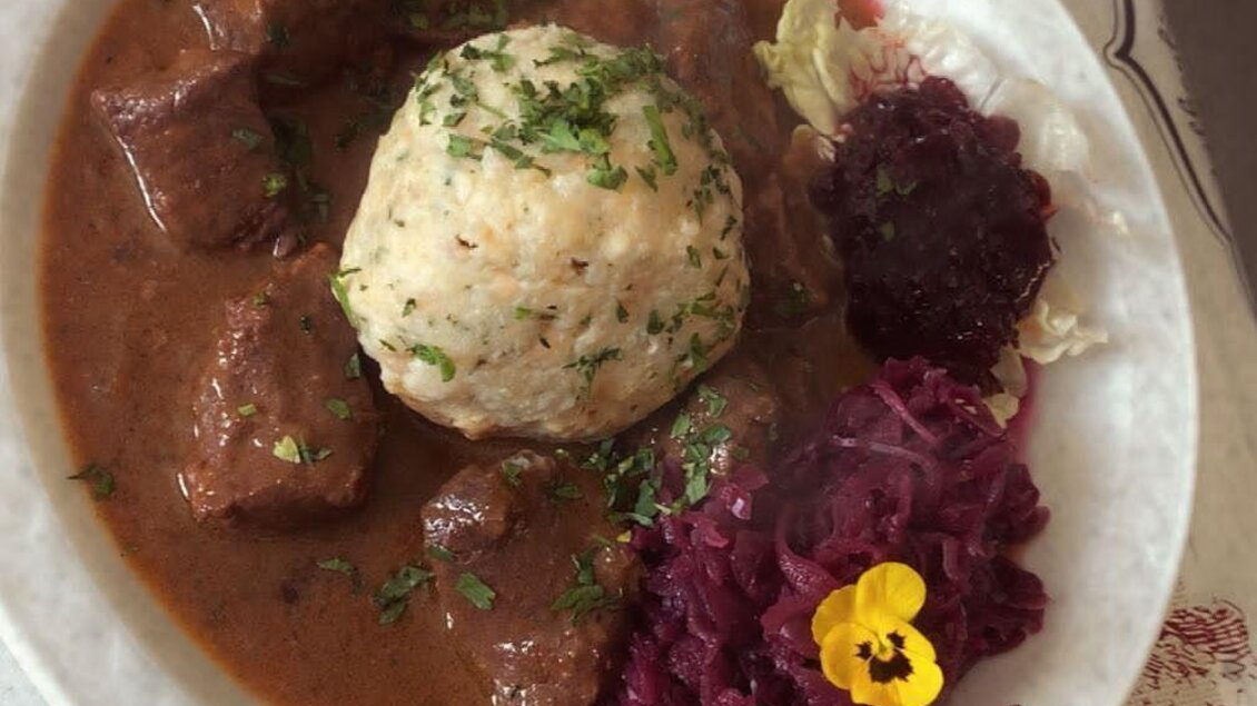 Ein Teller mit herzhaftem Rindfleisch, dazu Knödel und rotkraut. Das Gericht ist mit frischer Petersilie garniert und auf einem dekorativen Tisch präsentiert. | © Spechtenseehütte