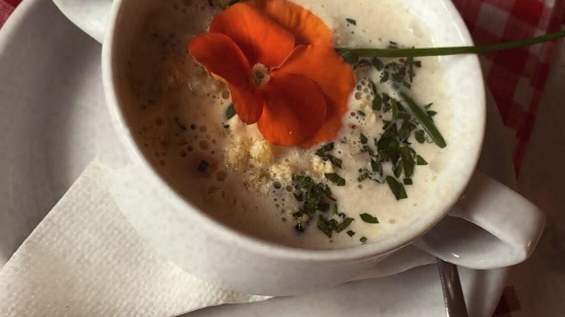 Eine Schüssel cremige Suppe mit einem Blütenblatt und frischen Kräutern. Der Untergrund ist mit kariertem Tischtuch in Rot und Weiß dekoriert. | © Spechtenseehütte