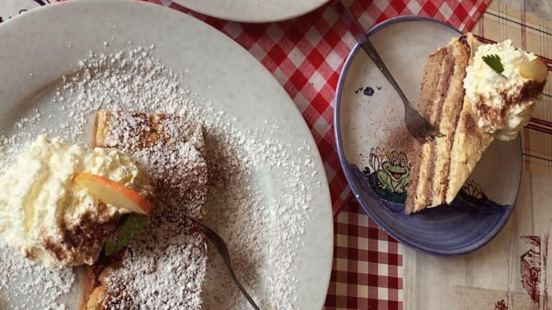 Zwei Teller mit Kuchen, garniert mit Sahne und Puderzucker, stehen auf einem rot-weißen Tisch. Neben den Tellern liegt ein weiteres Stück Kuchen auf einem kleinen Teller. | © Spechtenseehütte